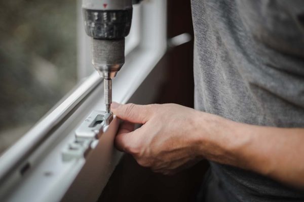 Young caucasian man installs metal fittings for plastic window frame with a drill, tightening a screw in a room where renovations are taking place, close-up side view with selective focus. The concept of home renovation, washing window frames.